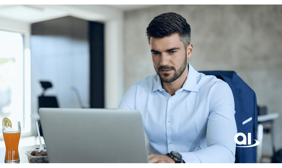 Man wearing a light blue shirt and sporting a short beard works on his laptop at a desk with orange juice and snacks.