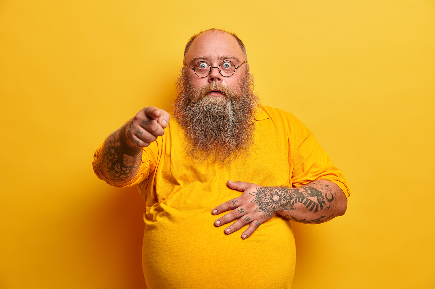 Stunned shocked bearded obese man points index finger at camera and holds tummy, reacts to overwhelming unexpected news, wears spectacles and yellow t shirt, poses indoor, feels impressed, excited